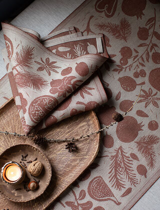 Tischläufer und Servietten in der Farbe Linen-Brown aus der Talven Tuoksut Kollektion, die aus 100% Leinen gefertigt und von Lapuan Kankurit aus Finnland stammt. Die Textilien sind ebenfalls mit weihnachtlichen Motiven wie Granatäpfeln, Tannenzweigen, Zapfen und Anis verziert. Neben den Leinenprodukten befindet sich eine Schale mit Walnüssen und einer Kerze, die eine warme und einladende Atmosphäre schafft. Die feinen Details der Motive und die hochwertige Verarbeitung sind deutlich erkennbar.