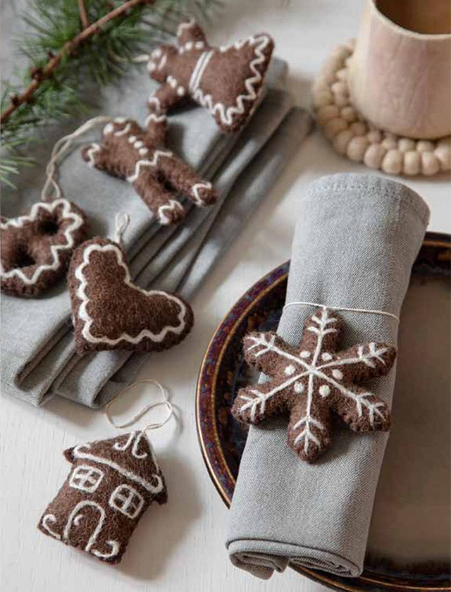 Die festliche Tischdekoration ist liebevoll für die Weihnachtszeit gestaltet und umfasst eine Auswahl an Wollfilz-Anhängern von Gry&Sif, die traditionellen Lebkuchenfiguren nachempfunden sind. Dargestellt sind ein Mann, eine Frau, ein Haus, eine Schneeflocke und ein Herz, alle detailreich mit weißen Linien verziert, die Zuckerguss imitieren. Diese Dekorelemente sind in einer rustikalen und gemütlichen Atmosphäre arrangiert, ergänzt durch einen grauen Stoffserviettenring mit Filzschneeflockenmotiv. Der Hintergrund wird durch weitere weihnachtliche Akzente wie Tannenzweige und eine hölzerne Perlenkette bereichert, was den Gesamteindruck sehr einladend und festlich macht.