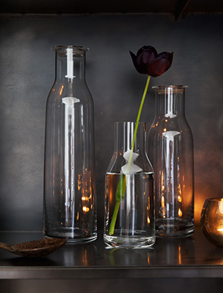3 Minima-Flaschen von Holmegaard als Blumenvasen mit einer schwarzen Tulpe. Dazu spiegelt sich das Licht von Teelichtern aus der Serie Cocoon, ebenfalls bei elbdal im Programm.