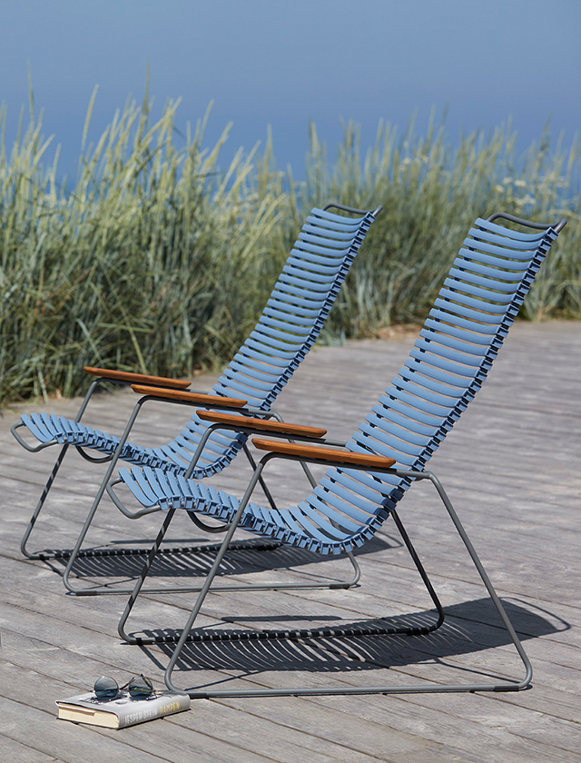 Zwei HOUE Click Lounge-Sessel in einem beruhigenden Taubenblau, die elegant auf einer Holzplattform an einem Strand platziert sind. Im Hintergrund weht Strandgras sanft im Wind, das Meer ist in der Ferne erahnbar. Die Sessel bestechen durch ihre schlanke, moderne Gestaltung mit lamellenartigen Sitz- und Rückenflächen, die auf einem robusten, grauen Metallgestell ruhen. Ergänzt wird das Design durch warme Holzarmlehnen, die einen natürlichen Kontrast zu den blauen Lamellen bilden. Ein Buch und eine Sonnenbrille liegen lässig vor den Sesseln auf dem Boden, was die Atmosphäre von entspanntem Genuss und Muße unterstreicht.