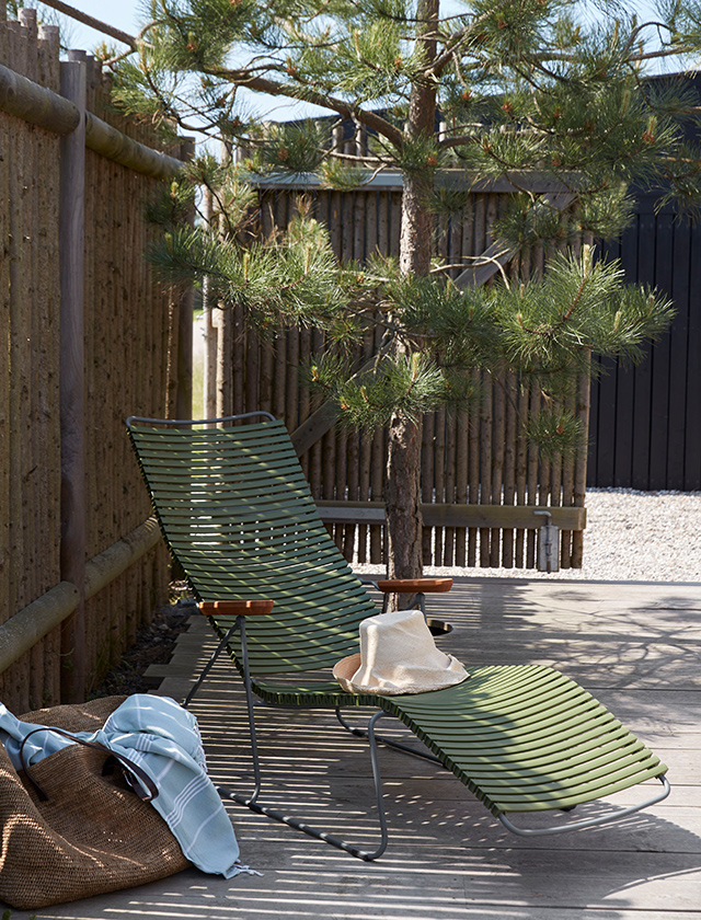Click Sonnenliege von HOUE, die in einem natürlichen, olivgrünen Farbton gehalten ist. Sie steht auf einer hölzernen Terrasse, umgeben von ruhiger Natur und einigen Kiefernbäumen, die einen Hauch von Waldesruhe vermitteln. Der Hintergrund wird durch einen Sichtschutzzaun aus Holz ergänzt, der das Bild eines privaten Rückzugsortes abrundet. Auf der Liege liegt entspannt ein Strohhut, der ein Gefühl von Sommer und Entspannung vermittelt. Neben der Liege befindet sich eine Tasche mit einem darin eingeklemmten blauen Tuch, was auf eine kürzlich genossene Auszeit hindeutet. Die Szene wirkt einladend und friedlich, ein idealer Ort zum Entspannen im Freien.