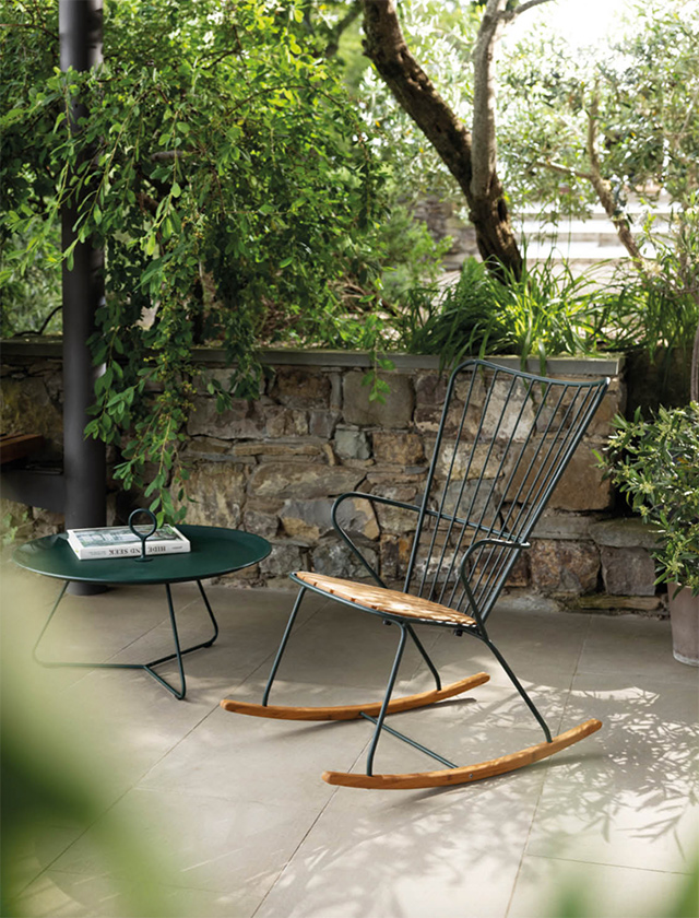 HOUE Paon Rocking Chair, einen Schaukelstuhl mit einem Gestell aus schwarzem Metall und Sitz- sowie Rückenfläche aus Bambus. Der Stuhl steht auf einer Terrasse, die von üppigem Grün und einer Steinmauer umgeben ist. Neben dem Schaukelstuhl befindet sich ein runder Eyelet Beistelltisch in passender dunkelgrüner Farbe, auf dem ein Buch und eine moderne, kreisförmige Sonnenbrille abgelegt sind. Diese Szene strahlt Ruhe und Entspannung in einer naturnahen Umgebung aus.