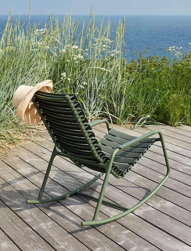 ReCLIPS Schaukelstuhl von HOUE in einem beruhigenden Olivgrün, der auf einer Holzplattform im Freien positioniert ist. Der Stuhl steht inmitten einer idyllischen Kulisse mit hohem Gras und wilden Blumen, die sanft im Wind wehen, mit dem ruhigen Blau des Meeres im Hintergrund. Ein Strohhut mit einer Schleife ist lässig über die Rückenlehne des Stuhls geworfen, was eine entspannte, sommerliche Atmosphäre schafft. Das Bild strahlt Ruhe aus und lädt dazu ein, sich zurückzulehnen und die Aussicht zu genießen.