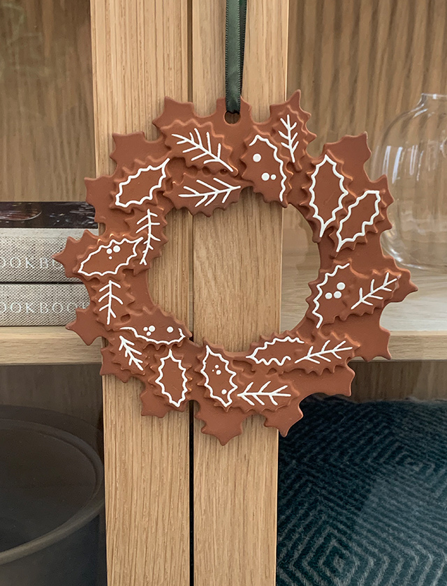 Gingerbread Kranz von Kähler Design, eine künstlerisch gestaltete Keramikarbeit, die traditionelle Weihnachtssymbole mit einem modernen Touch kombiniert. Der Kranz, geformt und bemalt, um wie ein klassischer Lebkuchen zu erscheinen, ist reich verziert mit weißem Zuckerguss-Design, das verschiedene winterliche Motive darstellt. Er hängt an einer schlichten, hölzernen Vitrine, was den warmen Terrakotta-Ton des Kranzes besonders hervorhebt.