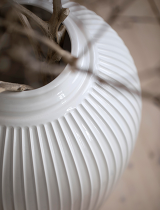 Detailaufnahme einer weißen Hammershøi-Bodenvase. Dargestellt ist der filigran geformte Vasenhals und die markanten Hammershøi-Rillen. Die Vase steht auf einem Holzfußboden und hält einen Strauß Zweige.