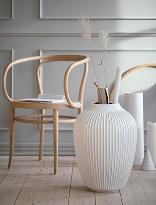 Das Bild zeigt eine weiße Hammershøi Bodenvase. Die große, stilvolle Vase wird hier zur Aufbewahrung von Papierrollen und Zeichnungen verwendet. Einige Trockengräser dienen der Verzierung. Den Hintergrund bilden der Stuhl 209 von Thonet und weitere grafisch-skulpturale Gegenstände, wie zum Beispiel ein Glasurkegel von Kähler Design.