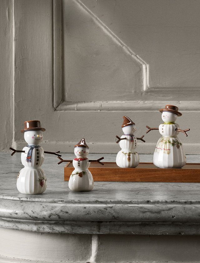 Liebevoll arrangierte Schneemann-Familie aus der Hammershøi Christmas Serie von Kähler Design, die aus glasiertem Porzellan gefertigt ist. Im Vordergrund links steht ein Schneemann mit einem braunen Zylinderhut und einem blauen Schal. Seine Arme, die aus kleinen Zweigen bestehen, und das freundliche Gesicht mit rosigen Wangen verleihen ihm einen charmanten Ausdruck. Der Körper ist mit dezenten winterlichen Motiven wie Zweigen oder Beeren geschmückt, was ihm eine elegante Note verleiht.