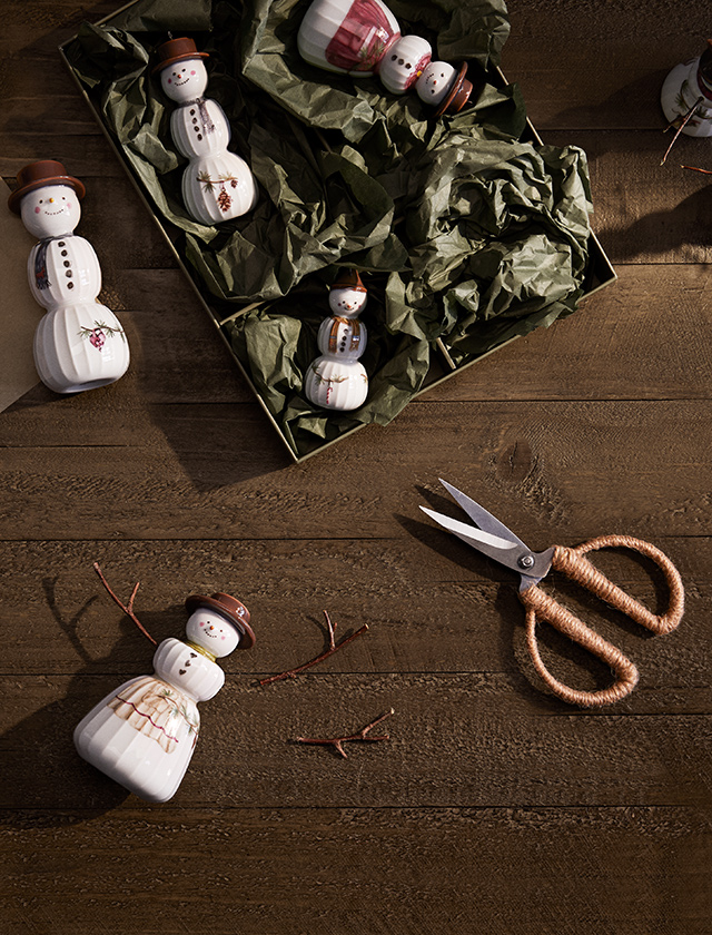 Schneemannfiguren der Hammershøi Weihnachtsserie von Kähler in einer dekorativen Szene arrangiert. Einige der Figuren liegen in einer Schachtel mit grüner Verpackungspapier, bereit zum Auspacken. Auf der Holztischoberfläche liegt eine Schere neben einer Schneemannfigur, die gerade mit kleinen Ästen als Arme dekoriert wird. Die handgemalten Details und die liebevoll gestalteten Accessoires der Schneemänner sind deutlich zu erkennen, und die gesamte Szene vermittelt eine gemütliche, weihnachtliche Stimmung.