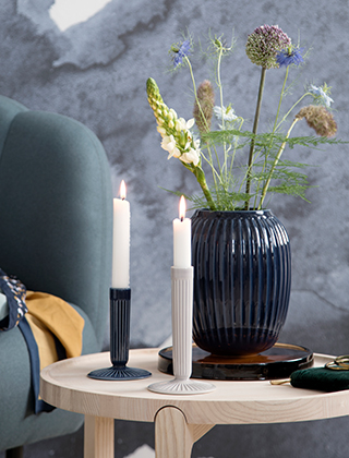 Ein stilvolles Arrangement aus zwei Hammershøi Kerzenleuchtern in Hellgrau und Indigoblau auf einem kleinen Tisch aus Eschenholz stehend. Neben den Leuchtern steht eine mittlere Hammershøi Vase in Indigoblau gefüllt mit frischgepflückten Blumen aus dem Garten.