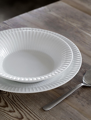 Das Bild zeigt zwei Teller und Besteck aus der Hammershøi-Serie von Kähler Design. Der tiefe Teller (Suppenteller) steht auf dem flachen Teller (Servierteller). Die weißen Teller mit den feinen Rillen sind aus Porzellan gefertigt und  stehen auf einem rustikalen Holztisch.