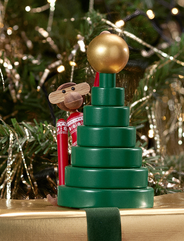 Kay Bojesen-Affen aus der Weihnachtskollektion neben einem grünen, stufenförmigen Weihnachtsbaum aus derselben Serie, gekrönt von einem glänzenden goldenen Ball. Der Baum, verfügbar in zwei Größen, und der Affe in einem rot-weißen Weihnachtspullover mit traditionellen Mustern, ergänzen sich in einer festlichen Szene. Im Hintergrund sorgen beleuchtete Girlanden für ein warmes, glitzerndes Ambiente. Diese Darstellung betont die handwerkliche Qualität und das ikonische Design von Kay Bojesen, bekannt für seine einzigartigen Sammlerstücke.