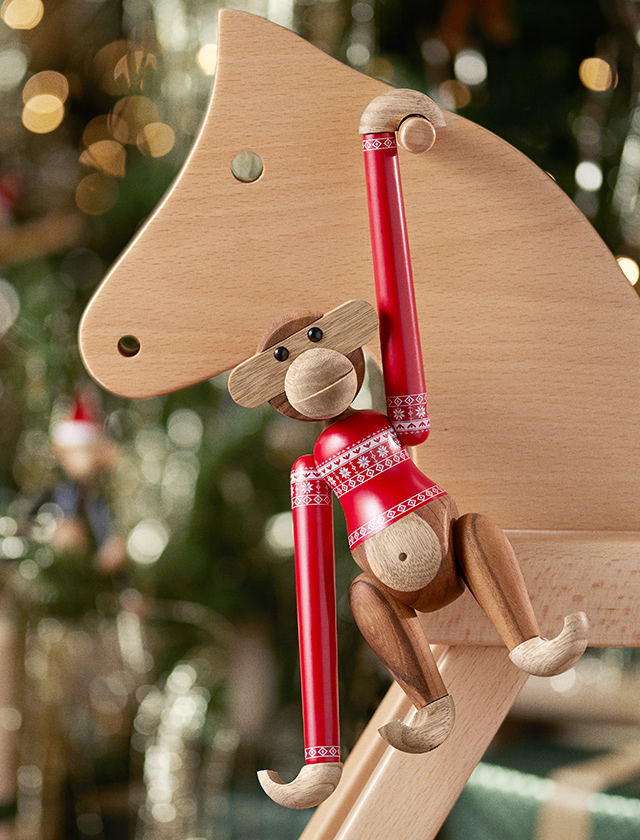 Kay Bojesen-Affen in festlicher Aufmachung, der auf einem hölzernen Schaukelpferd sitzt. Der Affe trägt einen auffälligen roten Weihnachtspullover, der mit einem traditionellen Muster aus weißen Sternen und Schneeflocken verziert ist. Die sorgfältig gedruckten Details des Pullovers, wie das Rhombusmuster an den Ärmelrändern, verleihen ihm ein besonders authentisches Aussehen. Die Szene spielt sich vor einem weihnachtlich geschmückten Hintergrund ab, der mit warmem Licht und glitzernden Dekorationen die festliche Stimmung unterstreicht. Dieses Ensemble aus hochwertigem Teakholz und liebevollen Details reflektiert das handwerkliche Geschick und den charakteristischen Humor von Kay Bojesens Design.