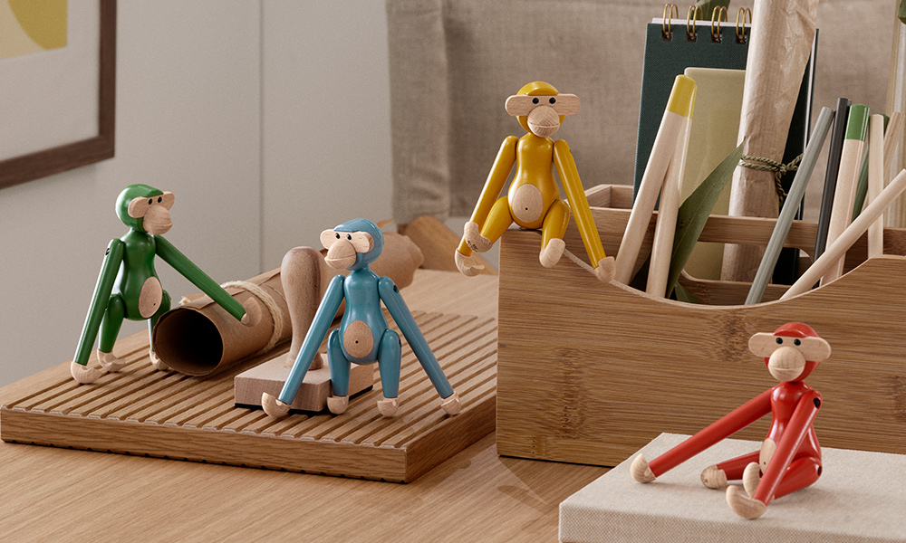 Mini-Affen aus der Vintage Kollektion von Kay Bojesen. Diese kleinen Holzfiguren, präzise gestaltet, sind in vier lebhaften Farben zu sehen: Grün, Blau, Gelb und Rot. Die Affen sind in verschiedenen Posen auf einem hölzernen Schreibtisch platziert, umgeben von kreativen Utensilien wie Zeichenrollen und Stiften in einem Holzbehälter.