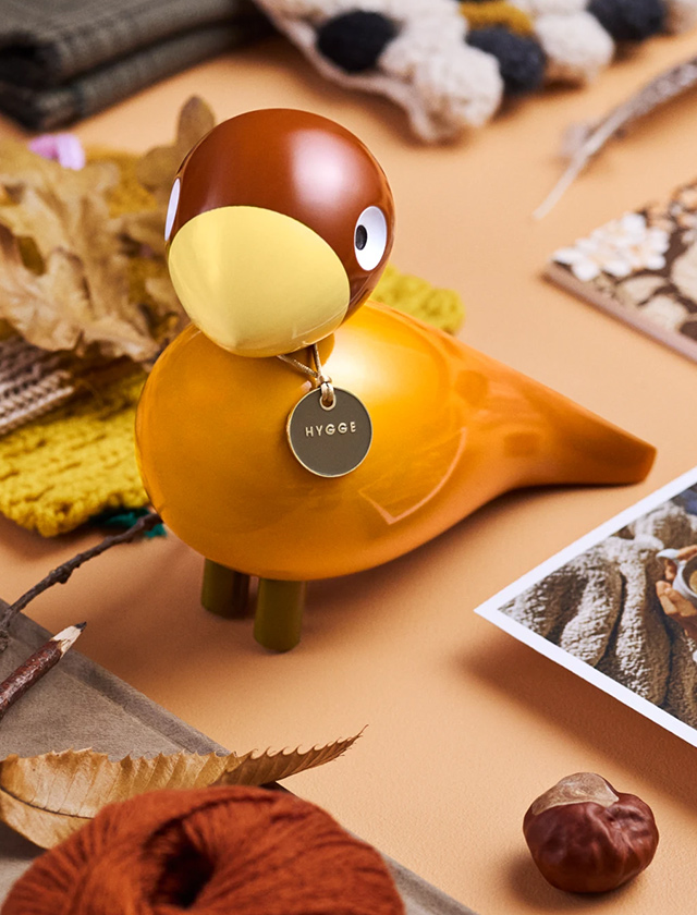 Orangegelbe Holzvogel-Figur „Hygge“ im skandinavischen Design auf herbstlichem Tisch-Setting mit Strick, Blättern und Kastanie
