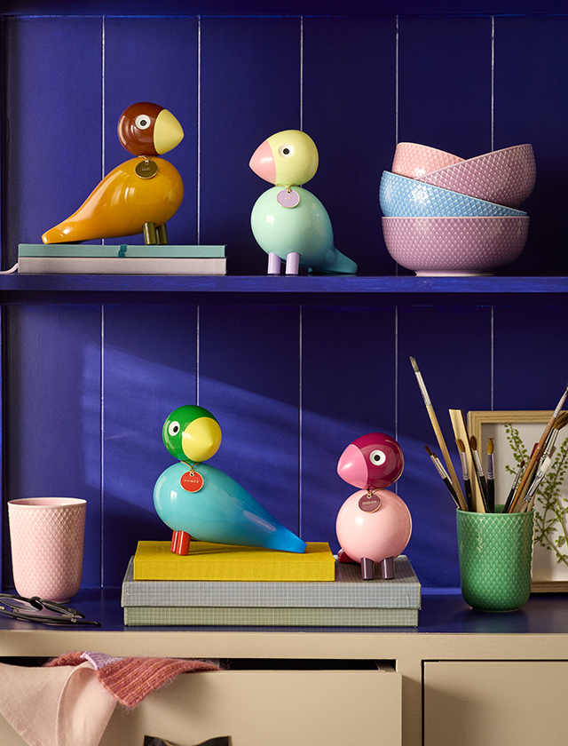 Vier farbenfrohe Kay Bojesen Story Birds aus Holz dekorativ auf einem Regal mit Büchern und Wohnaccessoires vor dunkelblauem Hintergrund arrangiert.