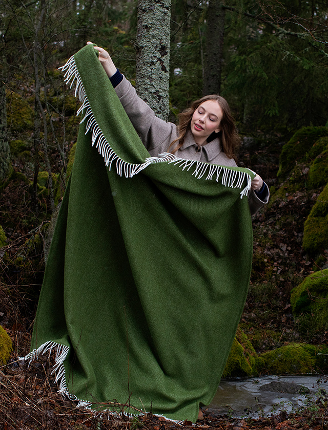 Arvo-Decke von Lapuan Kankurit aus Finnland. Die Decke besteht zu 100% aus Schurwolle und hat eine olivgrüne Farbe mit weißen Fransen an den Rändern. Die Person trägt einen hellen Mantel und steht in einem bewaldeten Gebiet, das von Moos bedeckten Steinen und Bäumen umgeben ist. Die Szene deutet auf eine kühle Jahreszeit hin, wahrscheinlich Herbst oder Frühling, da die Bäume keine Blätter haben und die Umgebung feucht erscheint.