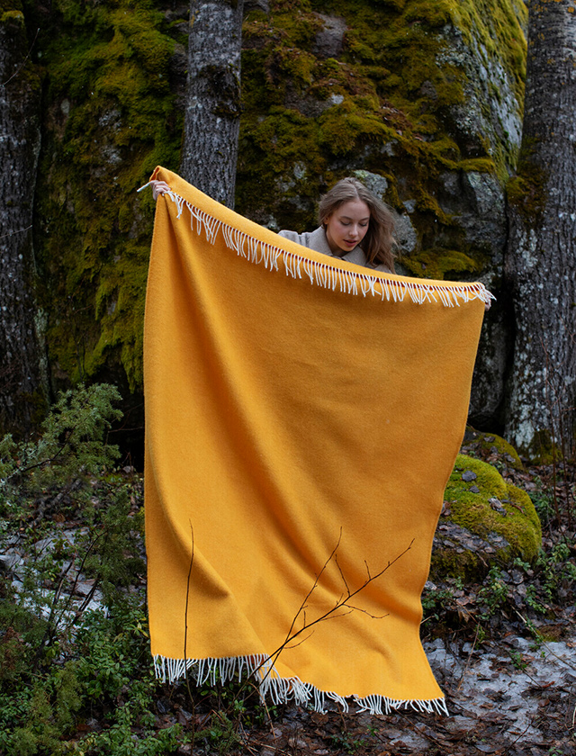 Arvo Decke von Lapuan Kankurit aus Finnland. Die Decke hat die Farbe White.Dyed-orange, was bedeutet, dass sie eine weiße Basis mit orangefarbenen Farbtönen aufweist und weiße Fransen an den Rändern hat. Die Person steht draußen in einem finnischen Wald, umgeben von einer natürlichen Umgebung. Im Hintergrund ist eine Felswand zu sehen, die mit Moos bewachsen ist, sowie mehrere Bäume. Der Boden zeigt Spuren von Schnee und ist mit verschiedenen Pflanzen bedeckt. Die Decke ist vollständig ausgebreitet und wird an den oberen Ecken gehalten. Die Szene strahlt eine ruhige und naturverbundene Atmosphäre aus.