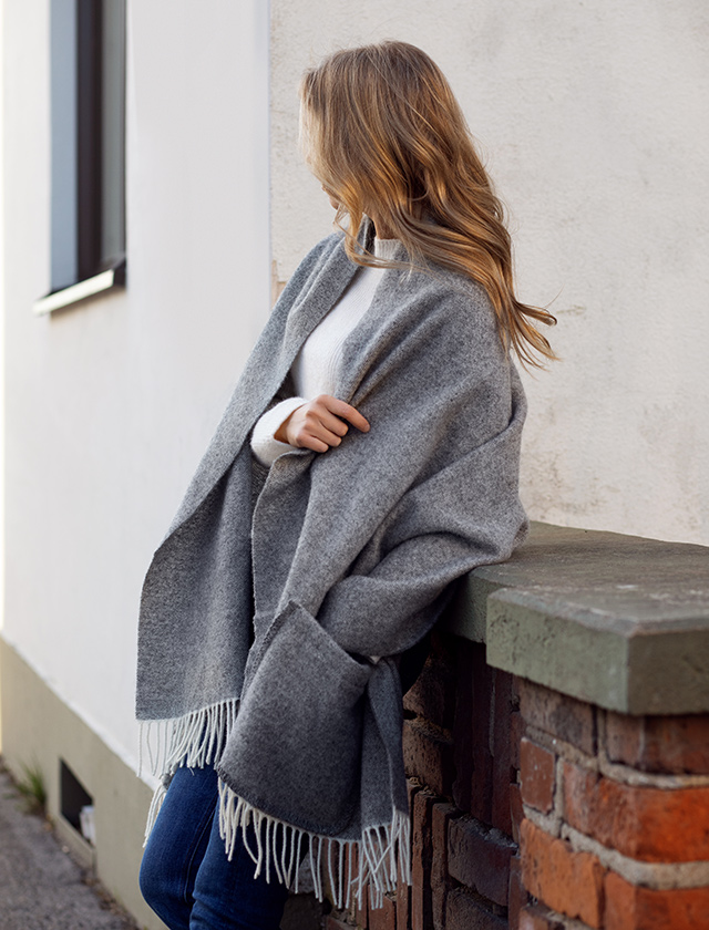 Lapuan Kankurit Arvo Pocket Shawl in Beige. Die Fransen an den Enden des Schals verleihen dem Outfit eine zusätzliche Note von Eleganz. Die neutralen Farben und die großzügige Größe des Schals sorgen dafür, dass er eine perfekte Wahl für kalte Tage ist, an denen sowohl Komfort als auch Stil gefragt sind. Die Position des Models und das natürliche Umgebungslicht vermitteln eine angenehme, entspannte Atmosphäre, ideal für Herbst- und Wintertage.