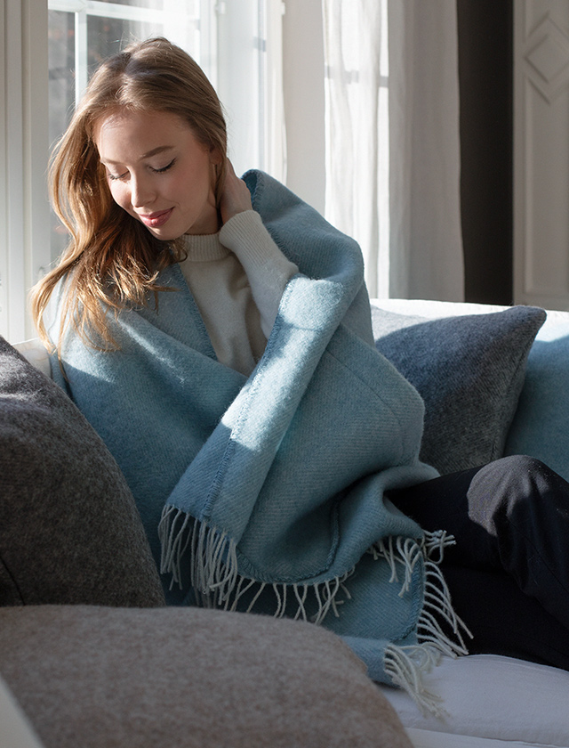 Der Arvo Pocket Shawl von Lapuan Kankurit in Dyed-Woad (Light-Blue) ist um eine Frau gewickelt, die auf einem Sofa sitzt. Sie lehnt sich entspannt zurück und genießt das Sonnenlicht, das durch das Fenster scheint. Der Schal hat weiße Fransen und ist in einer weichen, beruhigenden Farbe gehalten.