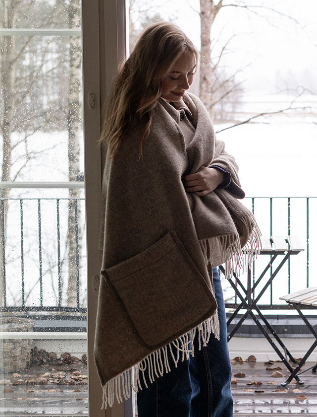 Der Arvo Pocket Shawl von Lapuan Kankurit in einem natürlichen Braunton ist um die Schultern einer jungen Frau gewickelt. Sie steht in einem Innenraum vor einer verglasten Tür mit Blick auf eine schneebedeckte Landschaft. Der Schal hat weiße Fransen und praktische Taschen, die gut sichtbar sind.