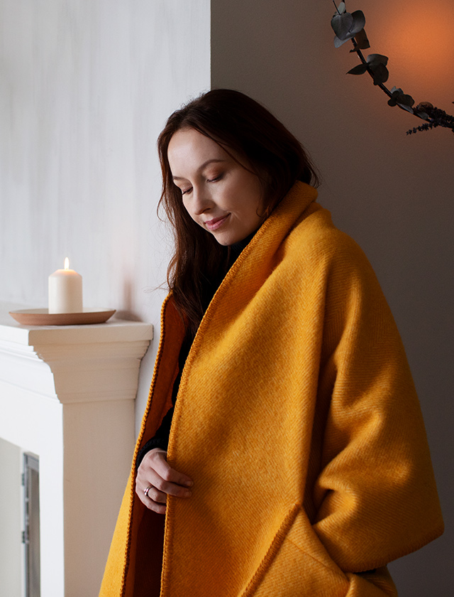 Der Arvo Pocket Shawl in leuchtendem Orange (White Coffe Dyed Orange) von Lapuan Kankurit ist um die Schultern einer Frau gewickelt, die vor einem Kamin steht. Der Schal hat eine intensive, warme Farbe und weiße Fransen. Die Szene wirkt gemütlich und warm, unterstrichen durch die Kerze auf dem Kaminsims.