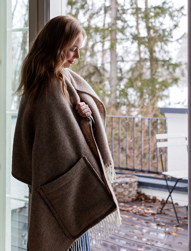 Der Arvo Pocket Shawl in einem natürlichen White Natures Brown von Lapuan Kankurit wird von einer Frau getragen, die auf einer überdachten Veranda steht. Der Schal hat weiße Fransen und praktische Taschen, die gut zur Geltung kommen. Die Umgebung ist herbstlich und vermittelt eine ruhige, besinnliche Atmosphäre.