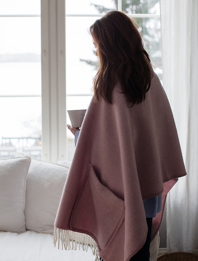 Eine Frau trägt den Arvo Pocket Shawl in White Rose von Lapuan Kankurit. Sie steht in einem gemütlichen Raum vor einem großen Fenster, das auf eine winterliche Landschaft blickt. Der Schal hat weiße Fransen und ist um ihre Schultern drapiert, während sie eine Tasse in der Hand hält.