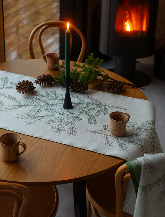 Eine gemütliche, winterliche Atmosphäre. Auf dem Tisch befindet sich ein weiß-grüner Havu Tischläufer von Lapuan Kankurit mit einem Tannenzweig-Muster. In der Mitte des Tischs steht ein schwarzer Kerzenhalter mit einer grünen Kerze, die brennt. Um die Kerze herum sind einige Tannenzapfen und ein paar grüne Zweige verteilt. Es stehen zwei braune Tassen auf dem Tisch, die handgemacht aussehen. Im Hintergrund ist ein Holzofen zu sehen, der eine warme und einladende Atmosphäre schafft. Die Stühle um den Tisch sind aus Holz und haben eine klassische, bogenförmige Lehne. Der Gesamteindruck ist rustikal und gemütlich, ideal für ein winterliches Beisammensein.