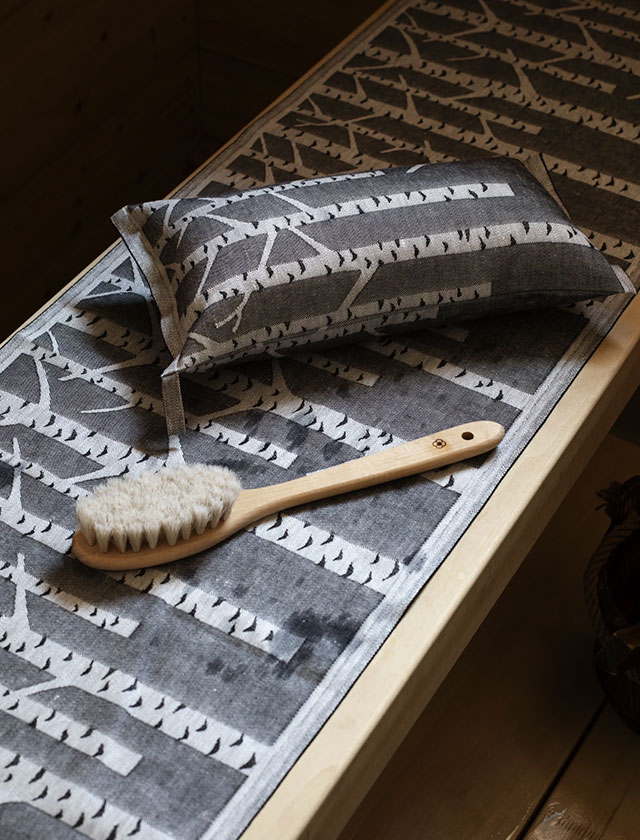 Stilvoller Komfort für deine Sauna: Das Bild zeigt das Saunakissen und die Sitzauflage Koivu von Lapuan Kankurit, gefertigt aus hochwertigem Leinen und verziert mit einem markanten Birken-Design in Schwarz-Weiß. Das grafische Muster, inspiriert von finnischen Birkenwäldern, schafft eine natürliche und beruhigende Atmosphäre. Ergänzt wird das Arrangement durch eine klassische Massagebürste, die das Saunaerlebnis abrundet. Die Textilien sind langlebig, saugfähig und atmungsaktiv – perfekt für nachhaltige Wellnessmomente im skandinavischen Stil.