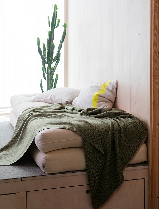 Gemütliches Schlafzimmer mit einer schlichten und modernen Einrichtung. Im Vordergrund liegt ein Bett, das mit einer beigen Matratze und mehreren weißen Kissen mit einem gelben Streifenmuster ausgestattet ist. Über das Bett ist eine Koli Merinowolldecke von Lapuan Kankurit aus Finnland in der Farbkombination Beige und Olive locker drapiert. Die Decke hat ein feines, dichtes Webmuster und vermittelt ein Gefühl von Wärme und Komfort. Im Hintergrund ist eine hohe grüne Zimmerpflanze, vermutlich ein Kaktus, zu sehen, die dem Raum eine frische und lebendige Note verleiht. Die Wände und der Rahmen des Bettes sind aus hellem Holz gefertigt, was dem Raum eine natürliche und einladende Atmosphäre verleiht.