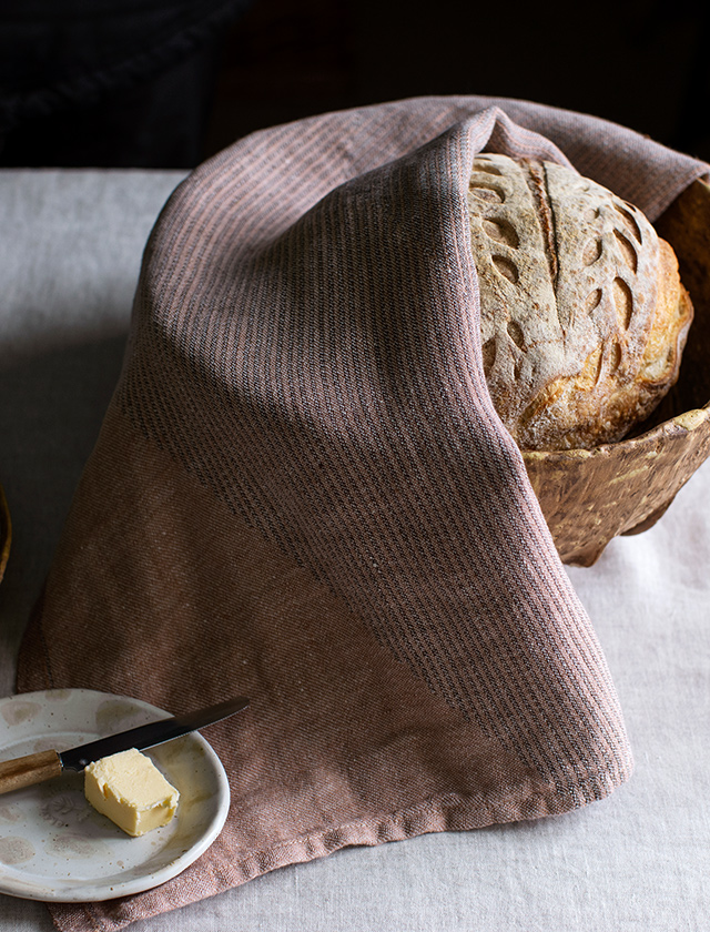 Rundes Brot, das teilweise mit einem Rinne Geschirrtuch abgedeckt ist. Das Geschirrtuch ist aus 100% Leinen und hat eine Brown-Cinnamon Farbe mit einem dezenten Streifenmuster. Das Brot hat eine schön gebackene, goldbraune Kruste mit kunstvollen Einschnitten. Das Geschirrtuch ist sorgfältig um das Brot gelegt und verleiht dem Bild eine gemütliche, rustikale Atmosphäre. Im Vordergrund befindet sich ein kleiner Teller mit einem Stück Butter und einem Buttermesser mit Holzgriff.