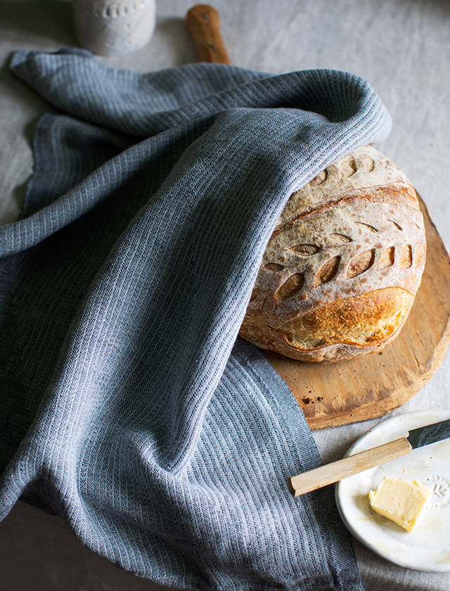 Rundes Brot, das auf einem Holzbrett liegt und teilweise mit einem Rinnen Geschirrtuch in Grtaphite-Grey bedeckt ist. Das Geschirrtuch ist aus 100% Leinen und hat eine graphitgraue Farbe mit einem feinen Streifenmuster. Das Brot hat eine knusprige, goldbraune Kruste mit dekorativen Einschnitten. Das Geschirrtuch ist elegant über das Brot drapiert. Im Vordergrund ist ein kleiner Teller mit einem Stück Butter und einem Buttermesser mit Holzgriff zu sehen, was dem Bild eine einladende und warme Stimmung verleiht.