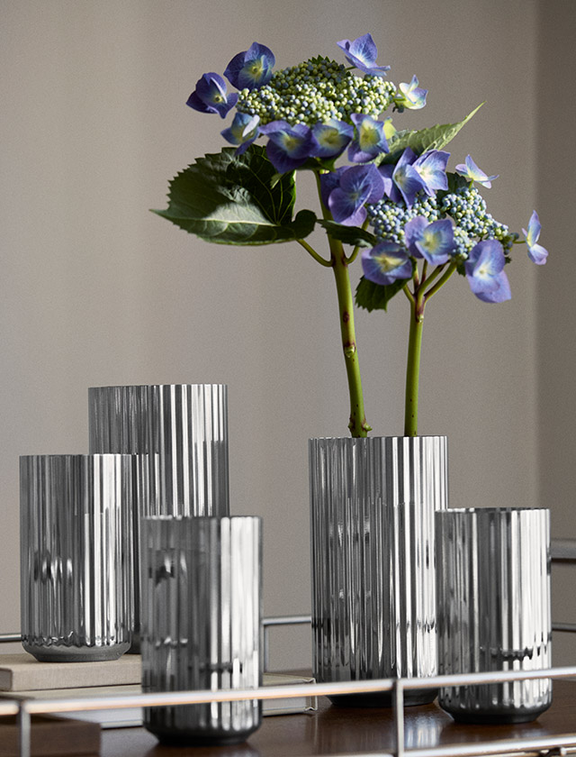 Lyngby Porcelæn Jubiläumsvase aus Edelstahl mit charakteristischer Rillenstruktur in verschiedenen Größen, dekoriert mit blauer Hortensie auf einem Design-Sideboard.