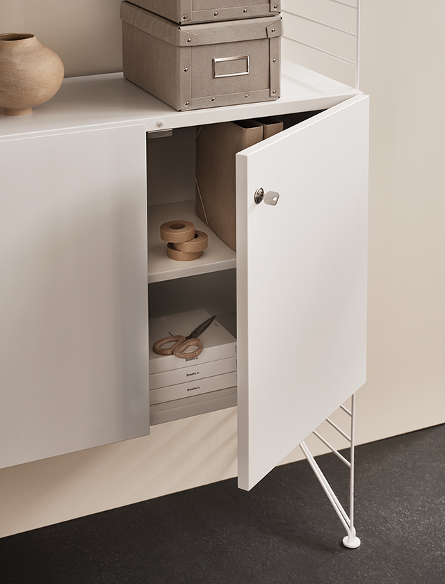 Eleganter weißer Schrank von String Furniture mit Klapptüren und einem Schloss. Dieser Schrank ist Teil des modularen und zeitlosen String Regalsystems, das sich durch minimalistisches skandinavisches Design auszeichnet. Der Schrank fügt sich nahtlos in moderne, funktionale Wohnräume ein. Auf der Oberseite des Schranks befinden sich dekorative Elemente und Stoffboxen in neutralen Farben, die zusätzlich Stauraum bieten. Im Inneren des Schranks sieht man praktische Ordnungshelfer wie Papierrollen und Scheren, was auf eine flexible Nutzung für Büro- oder Bastelmaterialien hinweist. Das Metallgestell verleiht Stabilität und unterstützt das klare, luftige Design, das typisch für die Marke String Furniture ist. Ideal für stilbewusste Kunden, die formschöne und funktionale Möbelstücke schätzen.